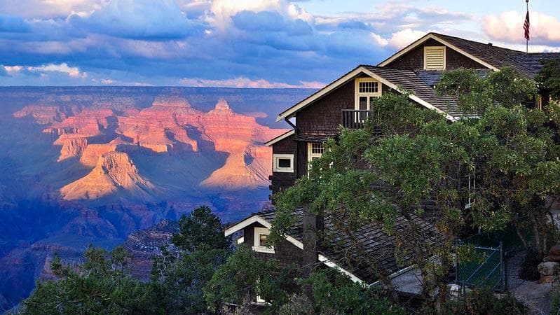 Wood frame building nestled by oak trees is overlooking sunset falling on colorful peaks and cliffs.