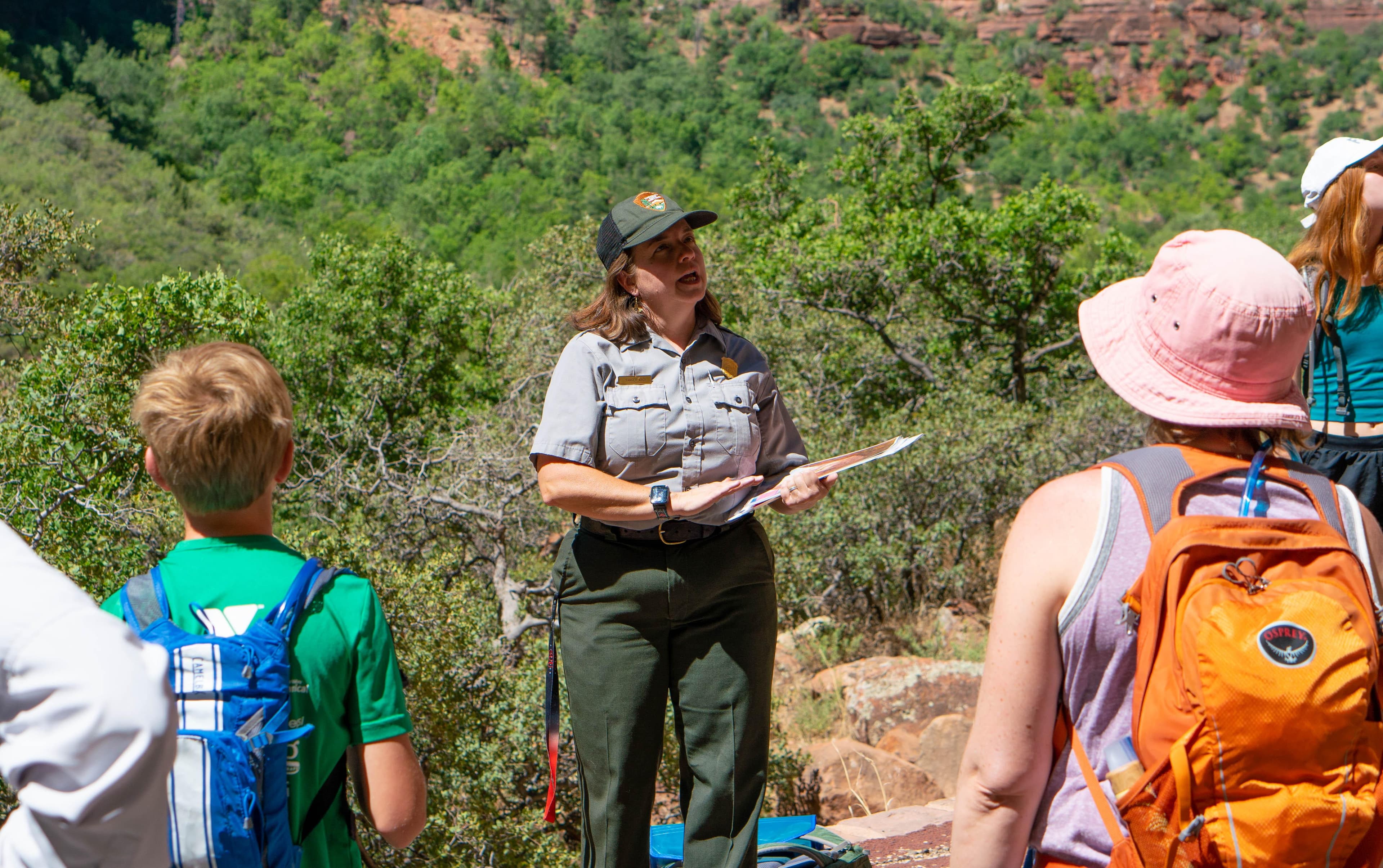 A park ranger speaks to a crowd of people circled around her on pavement.