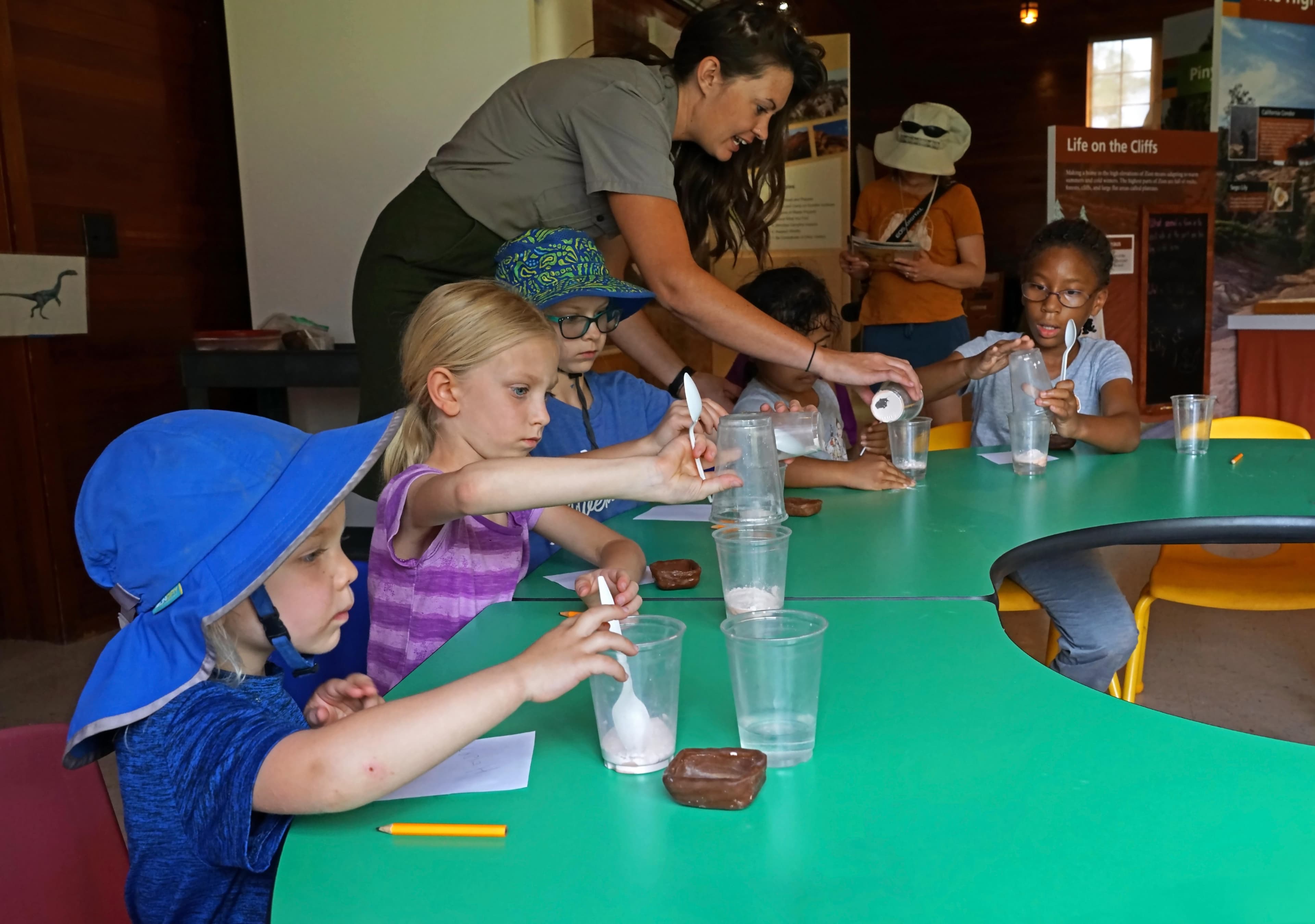 Four children seated at a curved green table mix together paste in plastic cups.