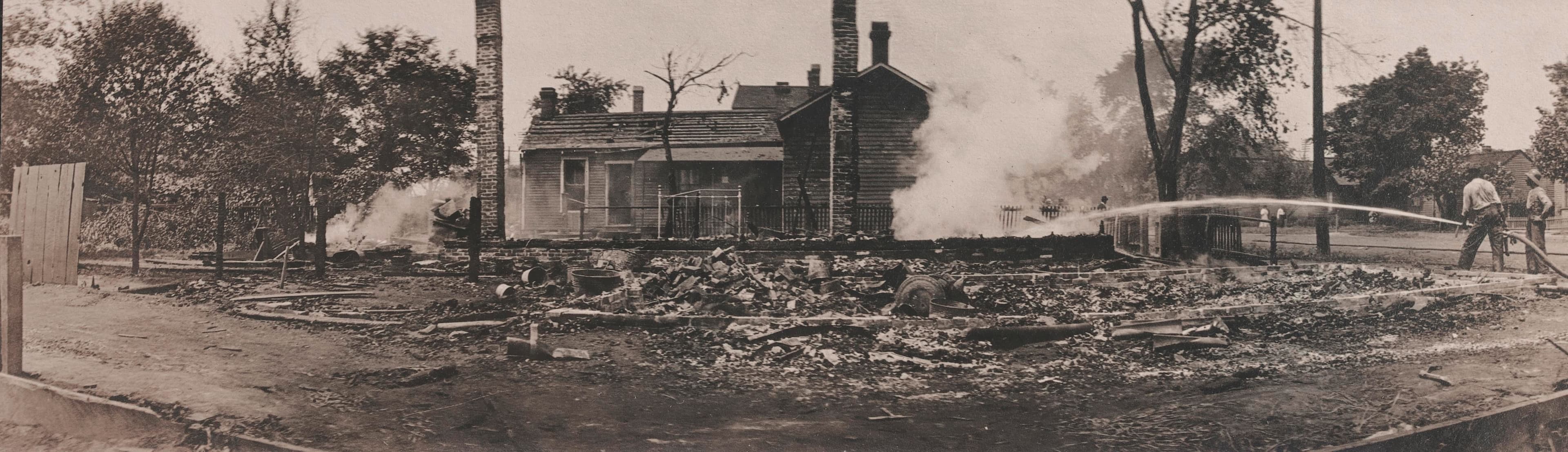 Historic photo of two people using a hose to extinguish a burning building.